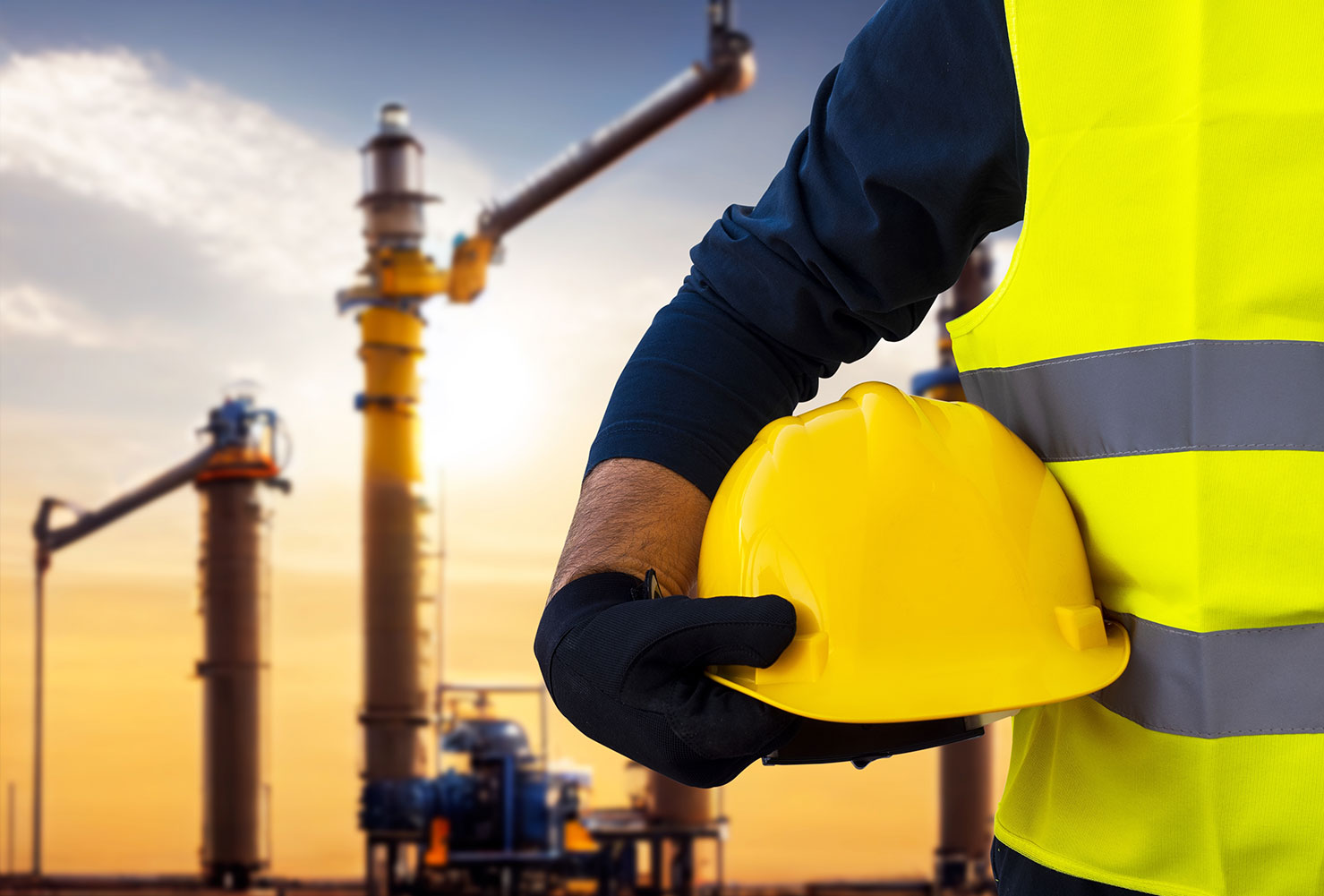 A close-up of an industrial worker wearing a high-visibility safety vest and black gloves, holding a yellow hard hat. In the background, there are industrial structures and pipes silhouetted against a golden sunset sky.