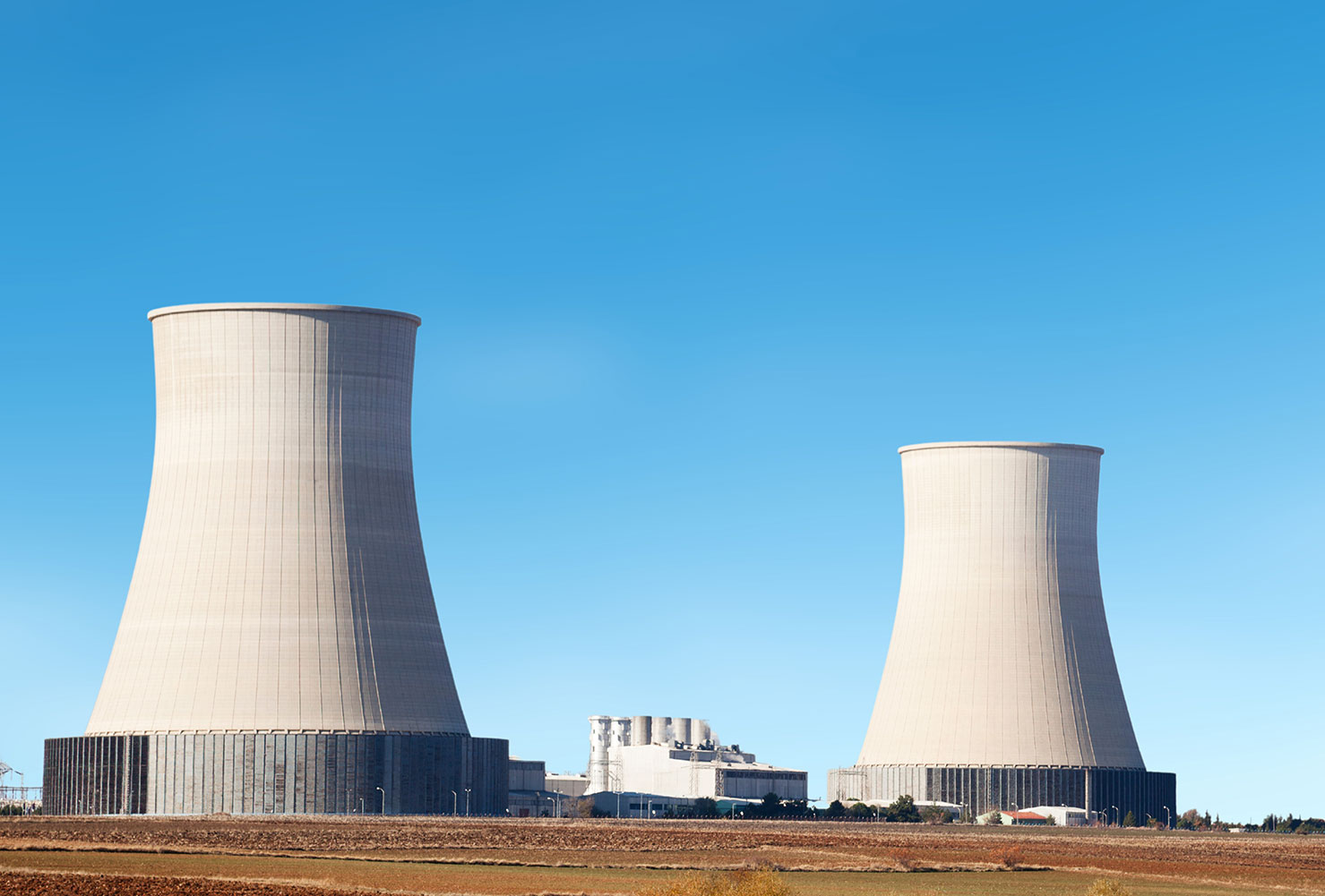 A large nuclear power plant with two prominent hyperboloid cooling towers rising into a clear blue sky. The surrounding area appears to be dry, flat land, and several industrial buildings are visible at the base of the towers.