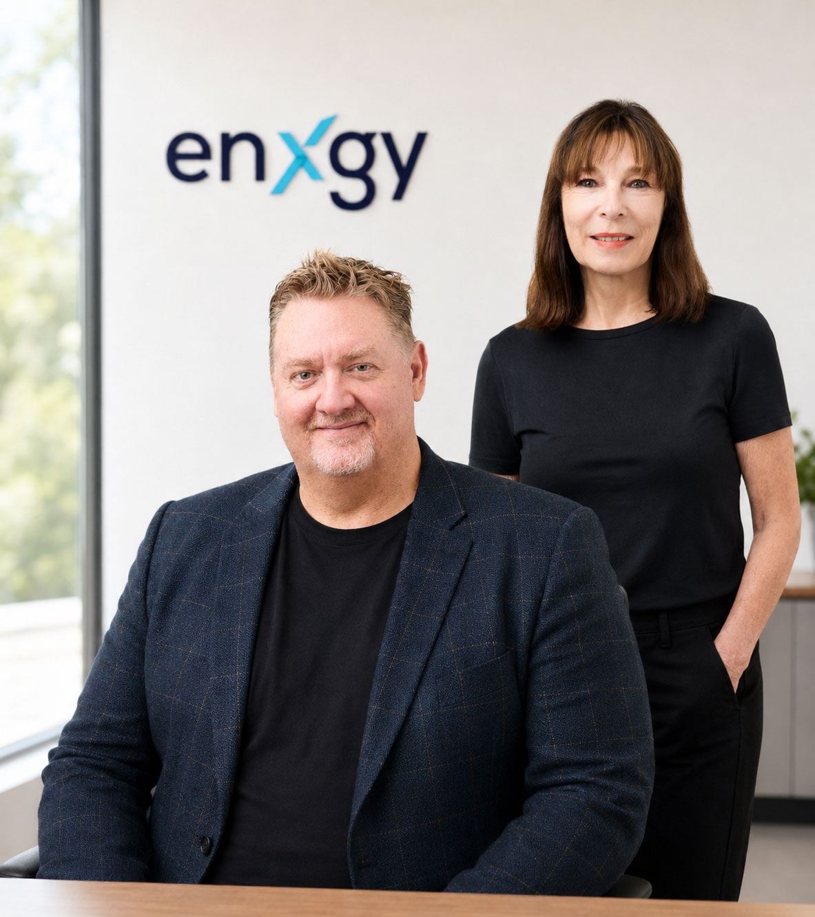 Steve sits in the foreground wearing a navy blazer, with Mel standing behind him in black, both facing the camera in a bright office setting with the Enxgy logo on the wall behind them.