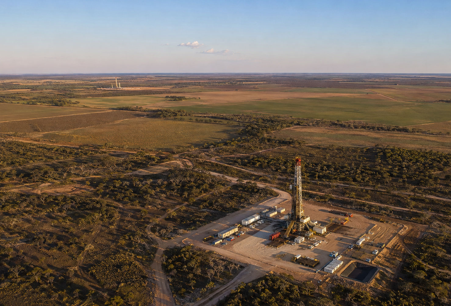 Aerial photograph of an oil and gas exploration well site in the flat agricultural landscape of Queensland's Western Downs, a drilling rig visible among low scrub and paddocks, a second distant well site visible on the horizon several kilometres away
